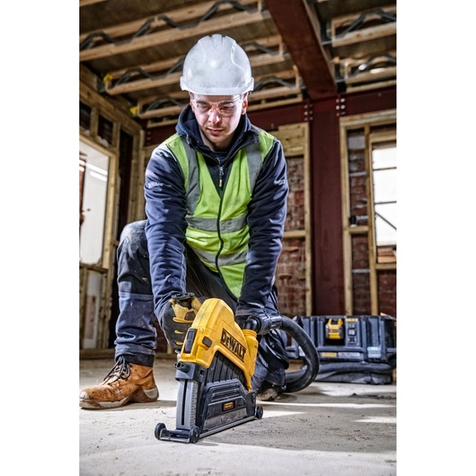 A person wearing a safety vest, hard hat, and work gloves uses a DEWALT power tool on a construction site floor. The tool is connected to a hose, and there is a DEWALT storage box in the background. The construction area has exposed beams and unfinished walls.