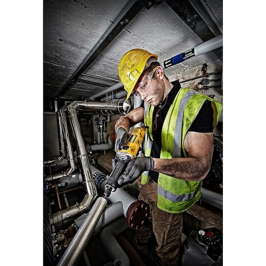 Worker uses DEWALT DWE4884 die grinder on metal pipe, wearing safety gear in an industrial setting.