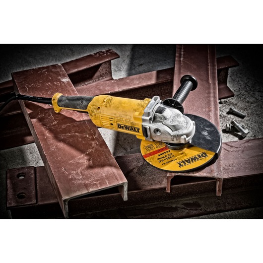 A DEWALT DWE490 angle grinder with a yellow body and a black handle is placed on metal beams. The grinder is plugged in and has a DEWALT-branded cutting disc attached. The setting appears to be a workshop or construction site with bolts lying nearby.