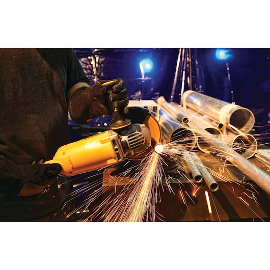 A person wearing protective gloves uses a yellow DEWALT angle grinder to cut metal pipes, producing bright sparks. Several metal pipes are visible on a workbench in an industrial setting.
