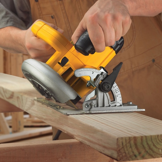 A close-up view of a person using a yellow DEWALT circular saw to cut a piece of wood at an angle. The saw features a metal base with measurement markings, and the person is holding the tool firmly with both hands.