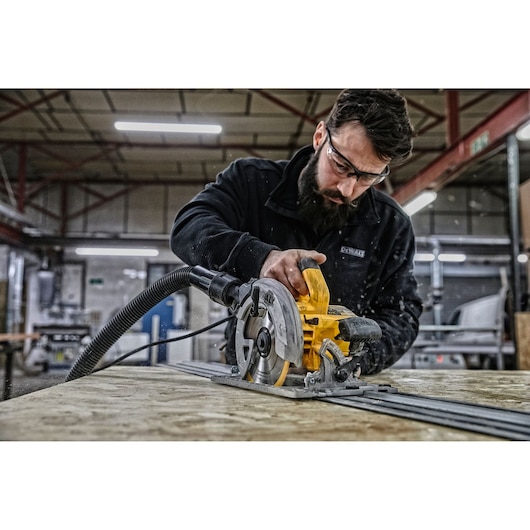 A person using a DEWALT circular saw to cut wood in an indoor workshop. The saw is yellow with a dust extraction hose attached. The person’s face is blurred.