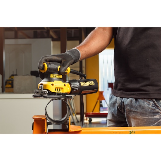 Close-up view of hands connecting a hose to a yellow DEWALT power sander, with a woodworking workspace in the background.