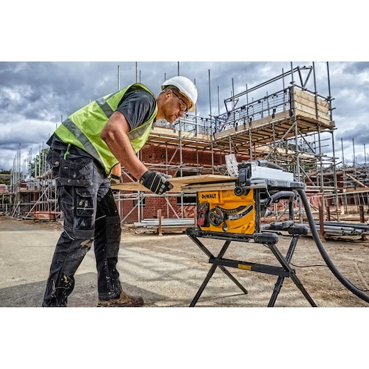 A worker wearing a safety vest and helmet uses a DEWALT DWE7485WS table saw to cut wood at a construction site. Scaffolding and unfinished buildings are visible in the background.
