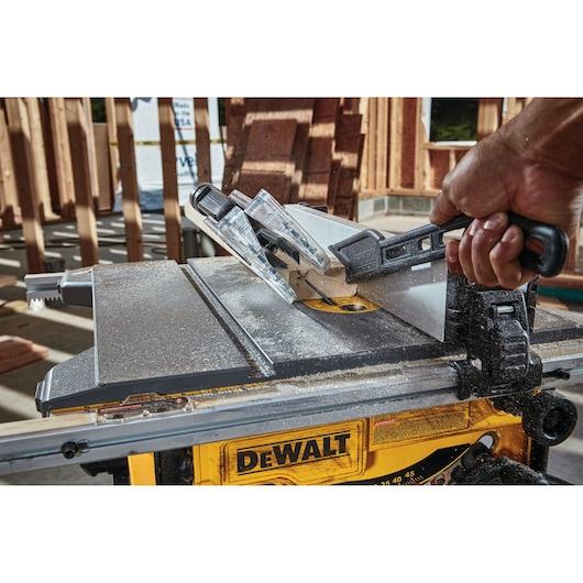 A close-up view of a DEWALT table saw in use, with a person's hand guiding a piece of wood through the cutting blade using a push stick. The saw is being operated at a construction site with wooden framing in the background.