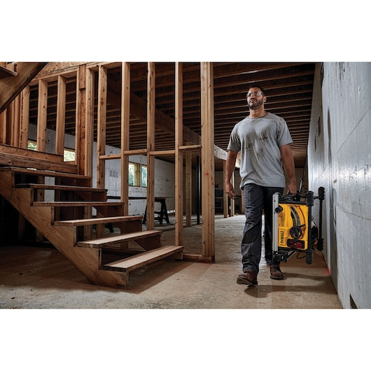 Person carrying a DEWALT portable table saw with stand in a partially constructed building, walking near unfinished wooden stairs and framed walls.