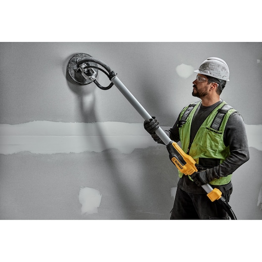 A construction worker wearing a hard hat and safety vest uses a DEWALT DWE7800 drywall sander with a telescopic handle to sand a gray wall.