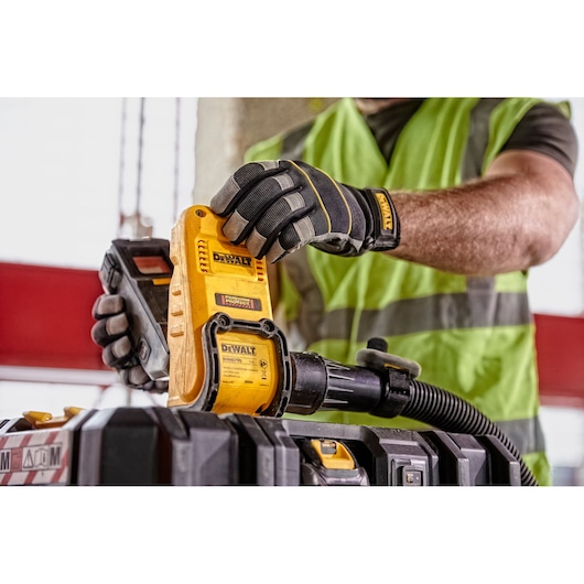 A person wearing protective gloves and a green safety vest is handling a yellow DEWALT device, which is connected to a hose and placed on a tool case in a construction setting.