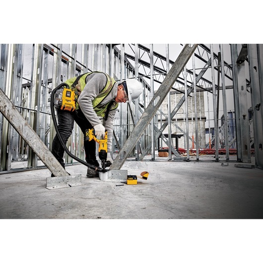 A construction worker wearing a safety helmet and vest uses a DEWALT power tool attached to a dust extractor to drill into a concrete floor at an industrial building site with metal framework.