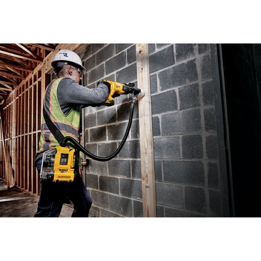 A construction worker wearing a helmet and safety vest uses a DEWALT cordless dust extractor system (DWH161D1) attached to a power tool while drilling into a concrete block wall at a construction site.