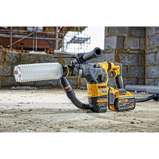 A DEWALT cordless rotary hammer drill with a dust extraction attachment, placed on a construction site floor with unfinished brick walls and scaffolding in the background.