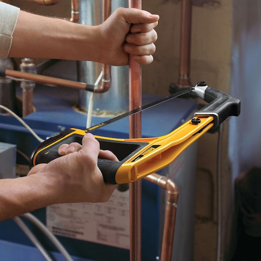 A person is using a yellow and black DEWALT hacksaw to cut a copper pipe in an indoor plumbing environment.