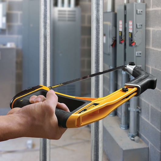 A person uses a yellow and black DEWALT hacksaw to cut a metal pipe in an industrial or utility room with electrical panels and exposed pipes in the background.