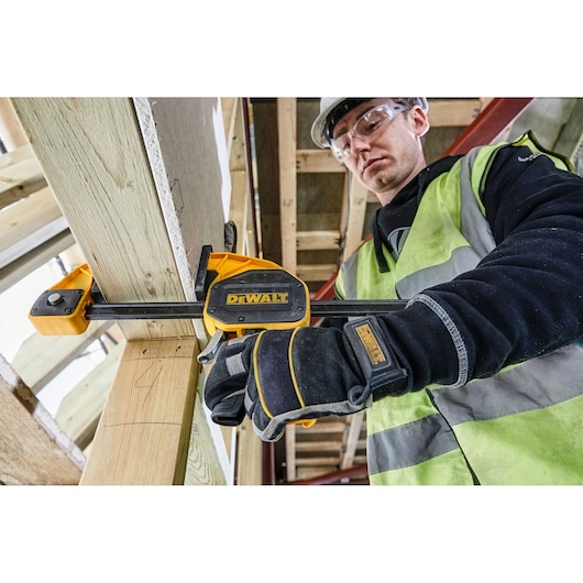 A gloved worker in a safety vest uses a DEWALT clamp to secure wooden beams at a construction site. The worker's face is blurred for privacy.