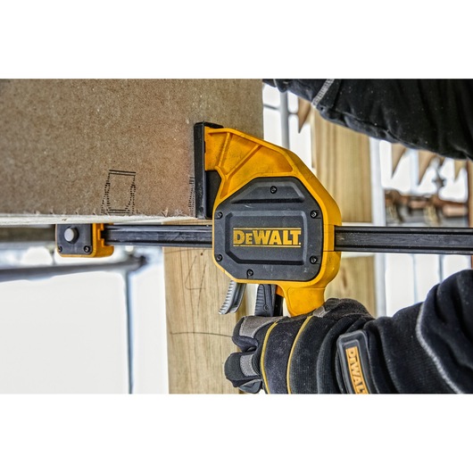 A DEWALT bar clamp is being used to secure a piece of wood in place. The clamp is yellow and black, and the user is wearing black work gloves.
