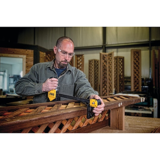 A person with a blurred face is using a DEWALT clamp to secure a piece of wooden lattice on a workbench in a workshop.