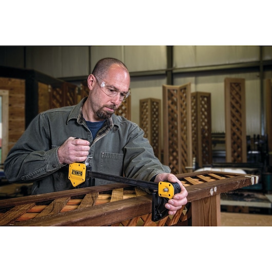 A person in a workshop using a yellow DEWALT corner clamp (SKU: DWHT0-83186) to hold wooden pieces together for assembly or repair.