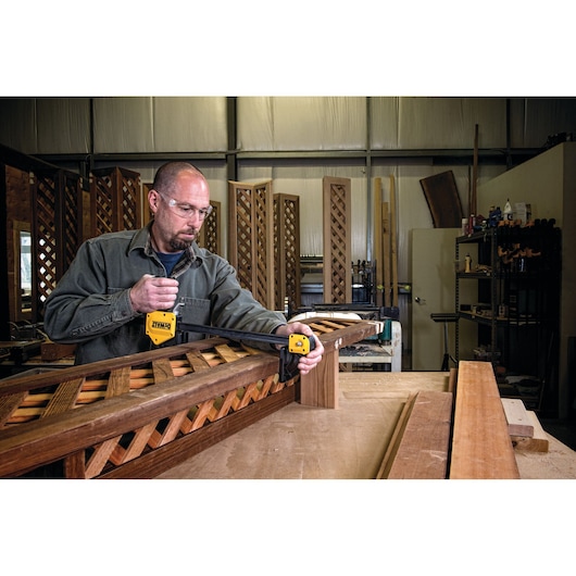 A person in a workshop is using a DEWALT clamp to secure a wooden lattice structure on a workbench. Various woodworking tools and materials are visible in the background.