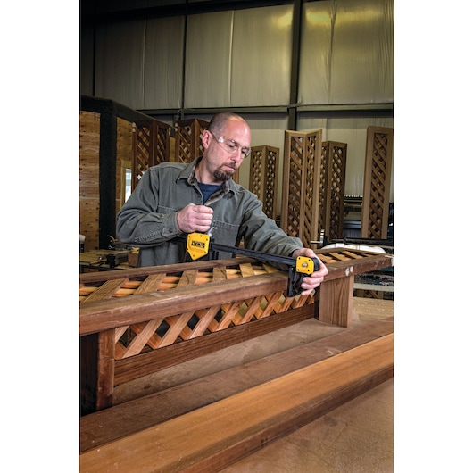 A person is using a DEWALT bar clamp to hold a wooden lattice structure in place on a workbench inside a workshop. The person's face is blurred for privacy.