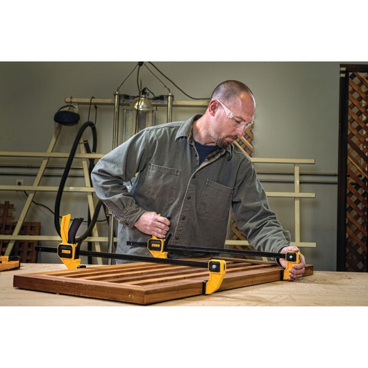 A person in a workshop uses two yellow DEWALT clamps to secure wooden boards on a workbench. The person's face is blurred and they are wearing a gray work shirt.