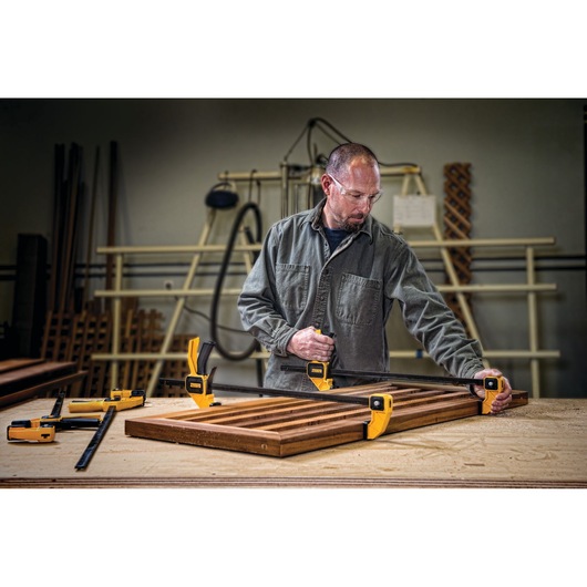 A person working in a woodworking shop uses DEWALT clamps to secure a wooden frame on a workbench. Additional DEWALT clamps are placed nearby. The person's face is intentionally blurred.