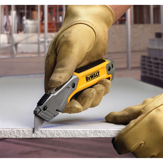 A person wearing work gloves uses a DEWALT utility knife to cut a piece of drywall or similar building material at a construction site.