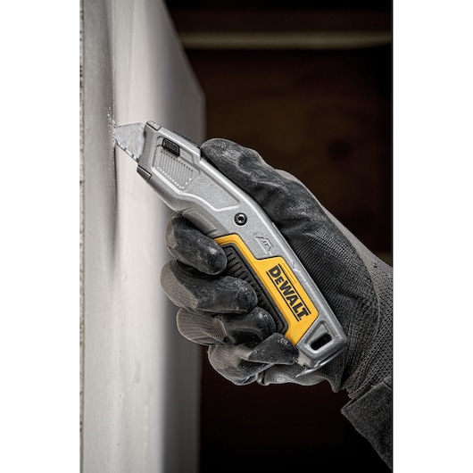 A gloved hand using a DEWALT utility knife to cut into a sheet of drywall.