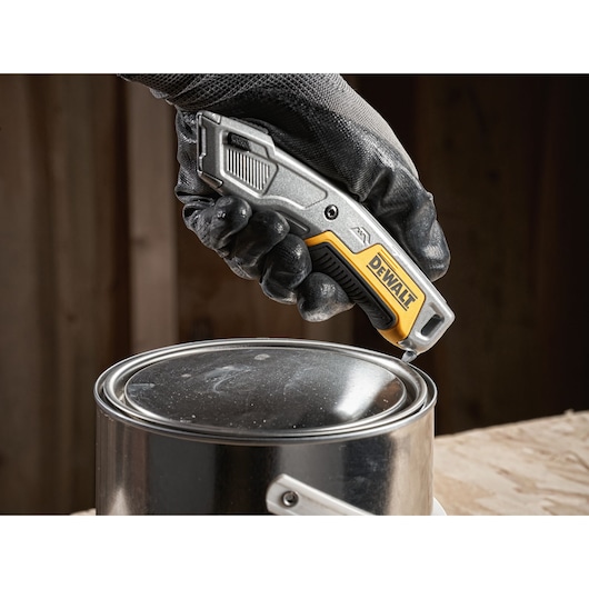 Gloved hand using a DEWALT utility knife to open a metal paint can on a wooden surface.