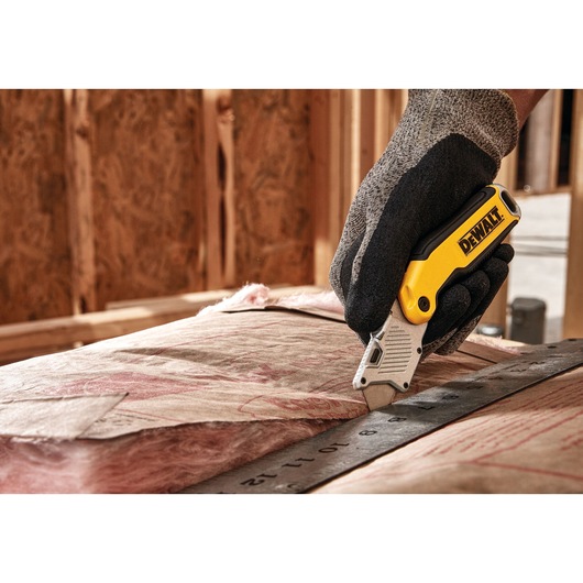 A gloved hand uses a DEWALT utility knife to cut insulation material along a metal ruler at a construction site.