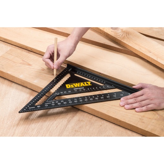 A DEWALT DWHT25228-0 photography square tool is placed on wooden boards. One hand holds the tool in position, while the other hand uses a pencil to mark measurements on the wood.
