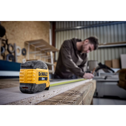 A DEWALT 8-meter tape measure is placed on a wooden workbench. In the background, a person with a blurred face is working, surrounded by tools and shelves in a workshop.