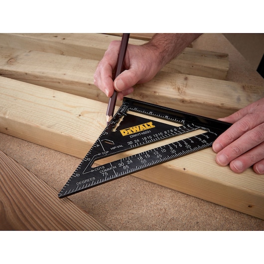 A person uses a DEWALT DWHT46031-0 black triangular rafter square to mark measurements on a piece of wood with a pencil. Several wooden planks are visible on a work surface.