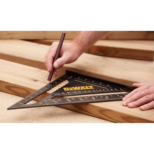 A person is using a DEWALT triangular carpenter's square to mark measurements on a wooden plank with a pencil. Several wooden planks are laid out on a work surface.