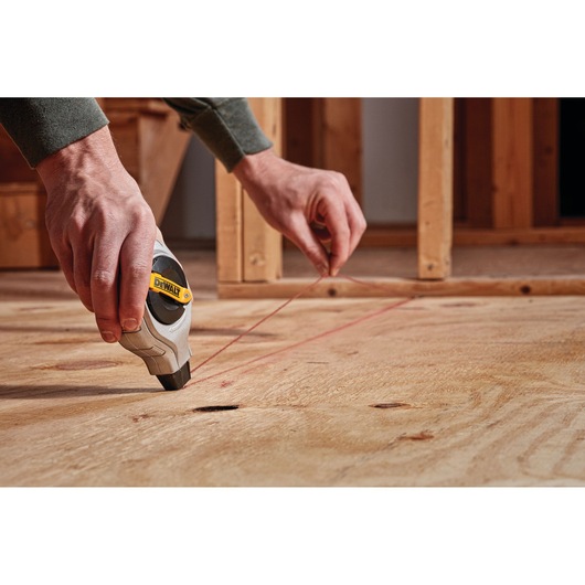 A person uses a DEWALT chalk line tool to mark a straight line on a wooden surface in a construction setting.