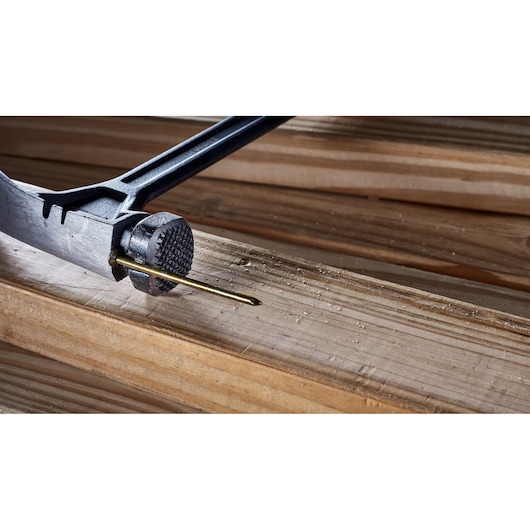 Close-up of a DEWALT hammer head with a nail being driven into a piece of wood, showing the textured striking surface and wooden planks in the background.