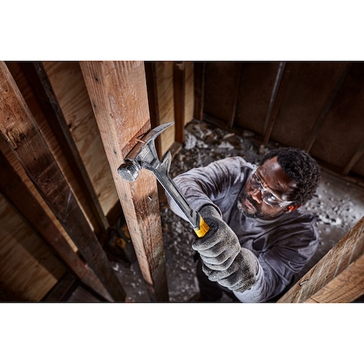 A person wearing gloves uses a DEWALT hammer with a yellow and black handle to strike a wooden beam inside a building under construction. The person's face is blurred.