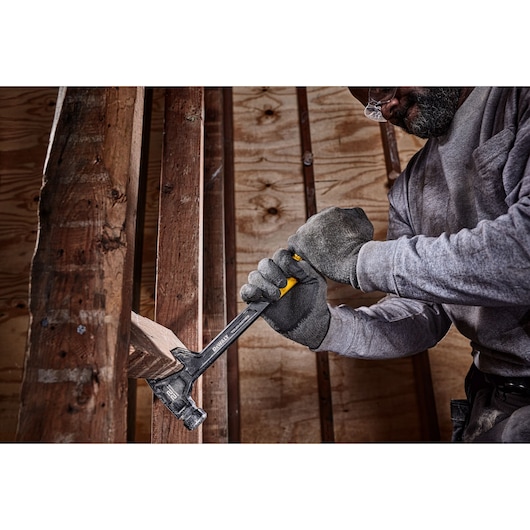A person wearing work gloves uses a DEWALT hammer to remove a wooden plank in a construction setting.