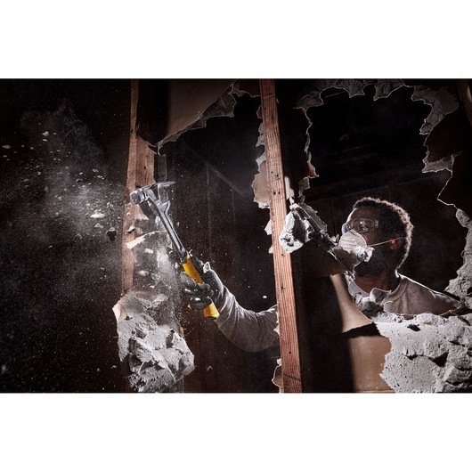 A person wearing gloves uses a DEWALT hammer to demolish a drywall, creating a cloud of dust and debris.
