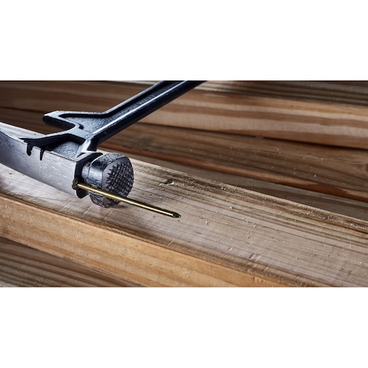 Close-up image of a DEWALT hammer using its textured striking surface to drive a nail into a wooden plank.