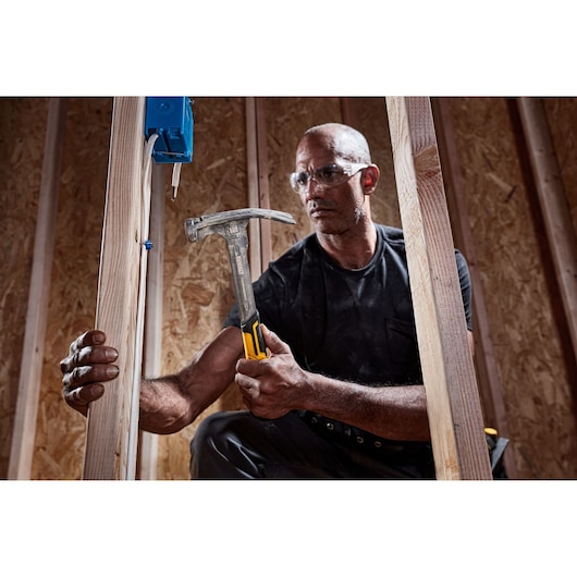A person holding a DEWALT claw hammer with a yellow and black handle, working on a wooden construction frame. Their face is blurred and they appear to be installing an electrical box onto wooden beams.