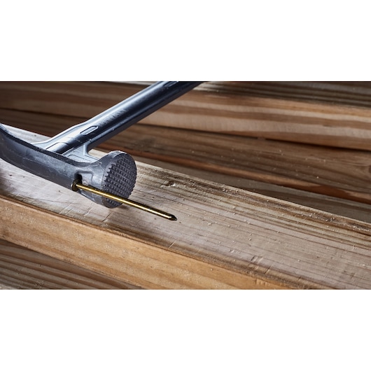 A close-up view of a DEWALT hammer driving a nail into a wooden plank. The hammer head and nail are clearly visible, with wood grain details in the background.