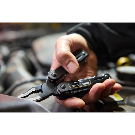 Hand holding a DEWALT black multi-tool with pliers, used for mechanical work in a workshop setting.