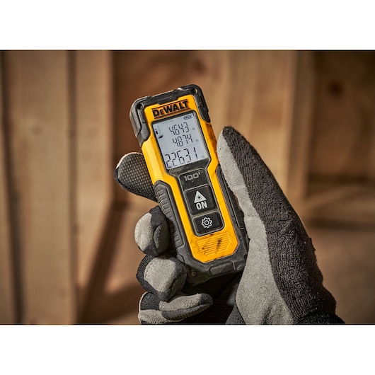 A gloved hand holding a DEWALT digital laser distance measurer, model DWHT77100, with a yellow and black casing and an illuminated display showing measurement readings. The background features wooden construction materials.