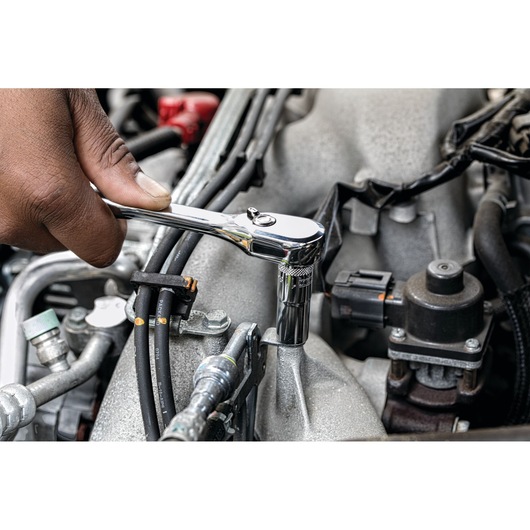 A hand using a chrome DEWALT ratchet wrench to tighten or loosen a bolt on a car engine, with various engine components and wires visible.