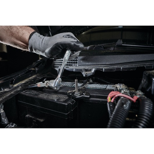 A gloved hand uses a DEWALT ratchet wrench to adjust a bolt on a car battery under the hood.