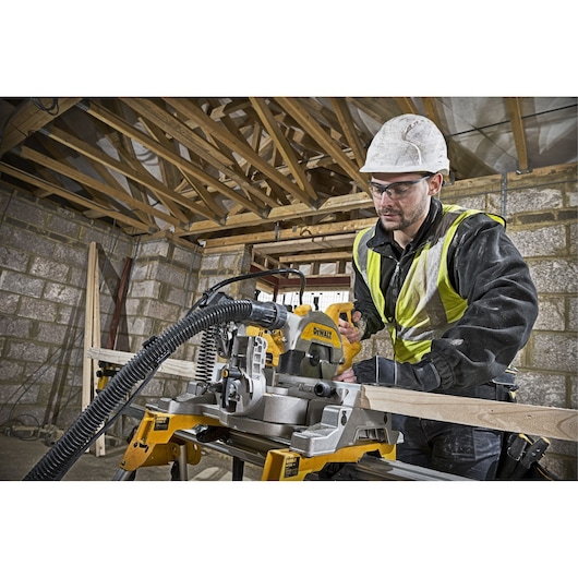 Person wearing a hard hat and high-visibility jacket operating a DEWALT DWS777 saw on a construction site, cutting a wooden plank.