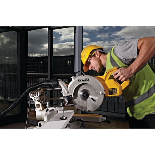 A worker wearing a safety helmet and high-visibility vest operates a DEWALT DWS778 sliding mitre saw, cutting a piece of material indoors. The background shows large windows and a partially cloudy sky.