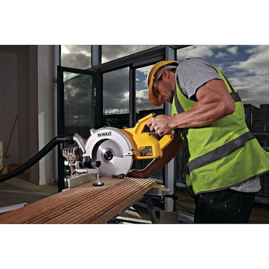 A person wearing a yellow safety helmet and a high-visibility vest operates a DEWALT circular saw to cut a piece of wood indoors.