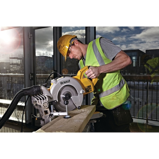 A person wearing a yellow hard hat and high-visibility vest is operating a DEWALT DWS778 mitre saw to cut a piece of wood in an indoor workshop with large windows.
