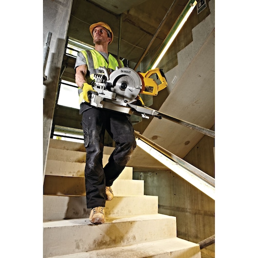 Person wearing a high-visibility vest, gloves, and a hard hat carrying a DEWALT DWS778 mitre saw down concrete stairs at a construction site.